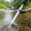 A person wearing black gloves uses a white handheld water sampler or pump device to collect water from a rocky stream, with green trees in the background.