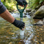 A person wearing black gloves inserts a white water sampler tube into a clear stream in a forest setting.