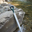 A white and dark green manual water filter straw with a loop handle, shown standing in shallow river water next to a large rock.