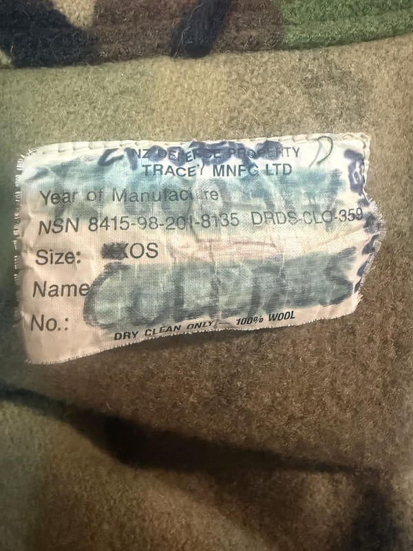 A close-up of a wool garment or fabric surface with a sewn-on care label. The label reads 'Tracey MNEC Ltd' and '100% Wool' in blue and black text on a beige/tan background.