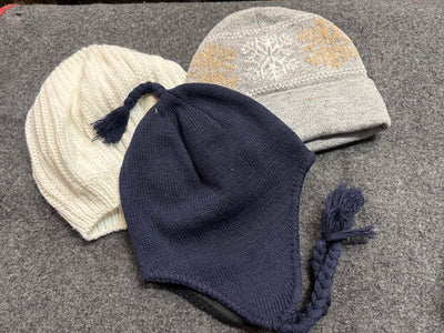 Three knit winter hats with ear flaps and tassels arranged on a gray textured surface: one cream, one navy blue, and one light gray with a gold snowflake pattern.