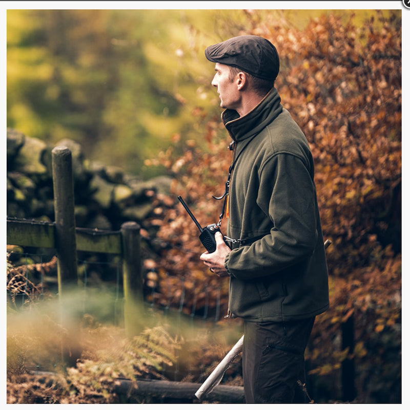 Man in a green jacket and cap holding a walkie-talkie in a forest setting