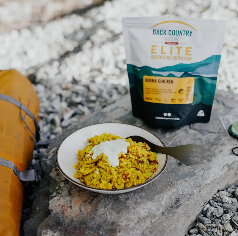 Campfire meal with a bowl of food and a 'Back Country Elite' package on a rocky surface.