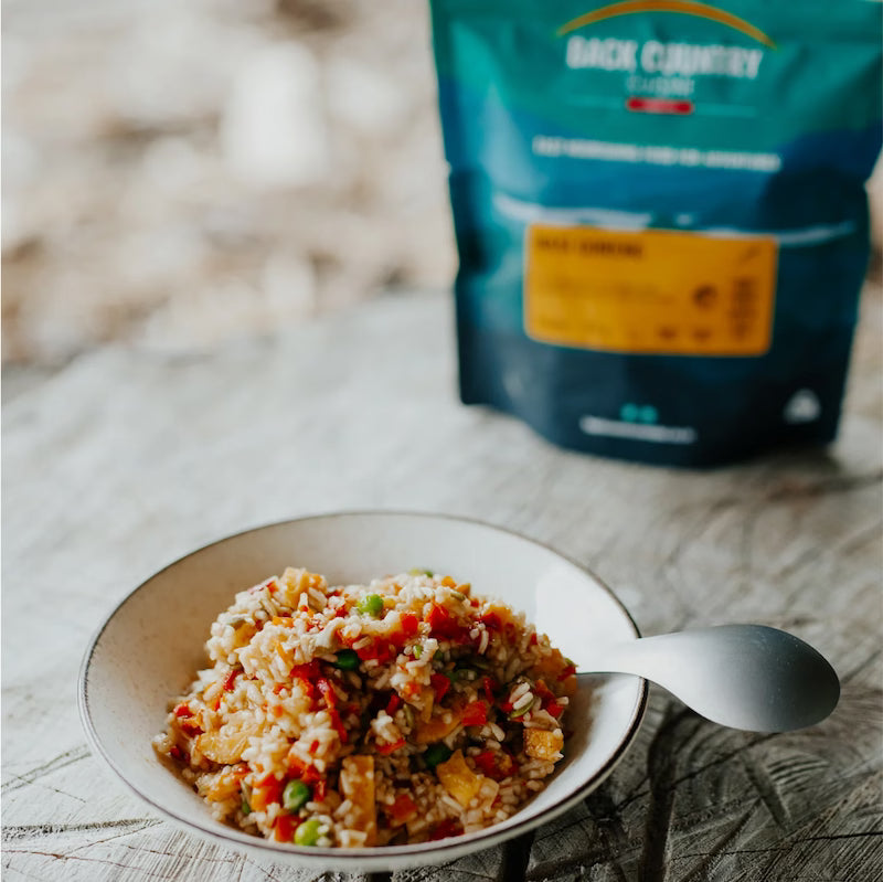  bowl of food with a spoon and a package of Back Country Cuisine in the background on a wooden surface