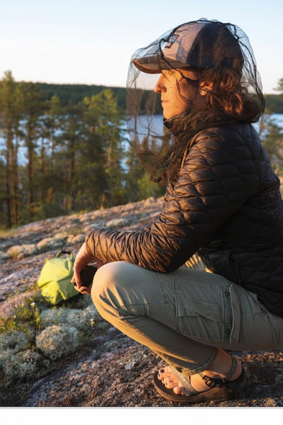 Person sitting outdoors by a lake, wearing a black jacket and cap with a net over their head.