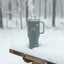 Gray insulated mug with steam on a snow-covered picnic table in a forest