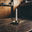 A single white taper candle in a small round metal holder, placed on a dark wooden table in a rustic room with wood-paneled walls and shelves.