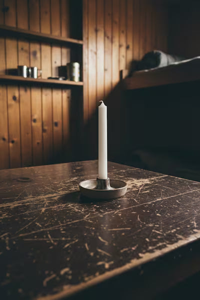A single white taper candle in a small round metal holder, placed on a dark wooden table in a rustic room with wood-paneled walls and shelves.