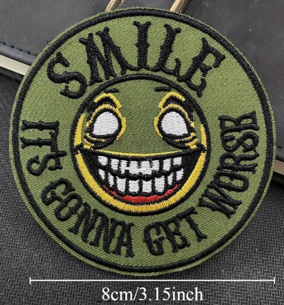 Embroidered patch with a smiley face and text 'SMILE IT'S GONNA GET WORSE' on a green background.
