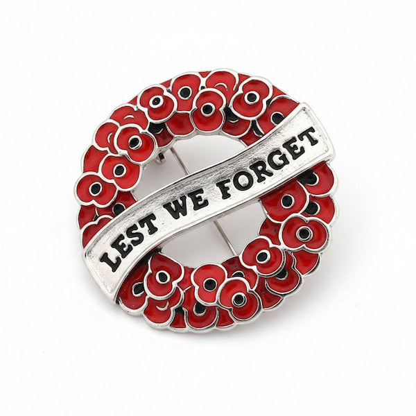 A circular silver-toned remembrance pin featuring red poppy flowers with black centers and a diagonal silver banner engraved with the words 'LEST WE FORGET'.