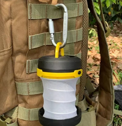 A tactical aluminum carabiner attached to a backpack with a collapsible silicone lantern in yellow and black, set against an outdoor background.