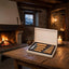 An open wooden presentation box holding a set of knives with light wood handles and silver blades, placed on a rustic wooden table in a warm, stone-walled room with a fireplace and candles.