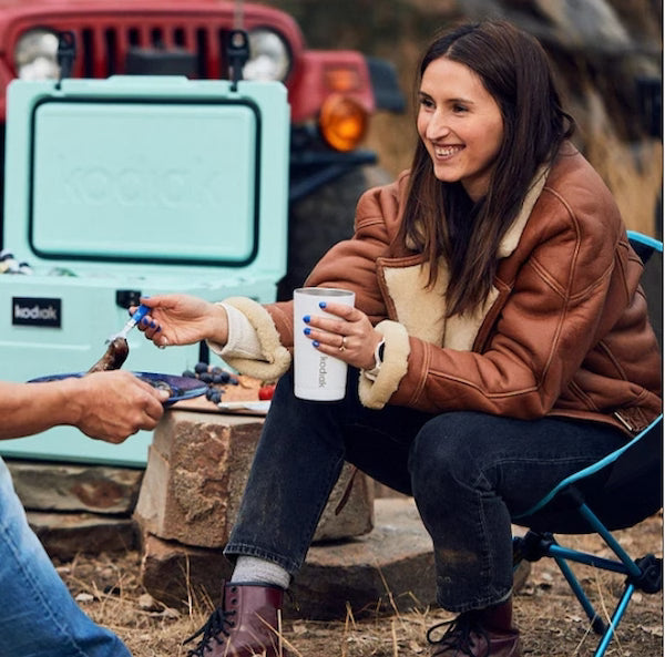Woman in a brown jacket holding a white mug outdoors with a teal cooler and red vehicle in the background.