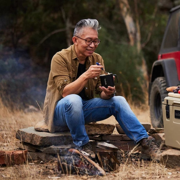 Man sitting outdoors by a vehicle, holding a mug and a pen.