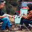 Two people sitting outdoors by a campfire, with a teal cooler and red vehicle in the background.