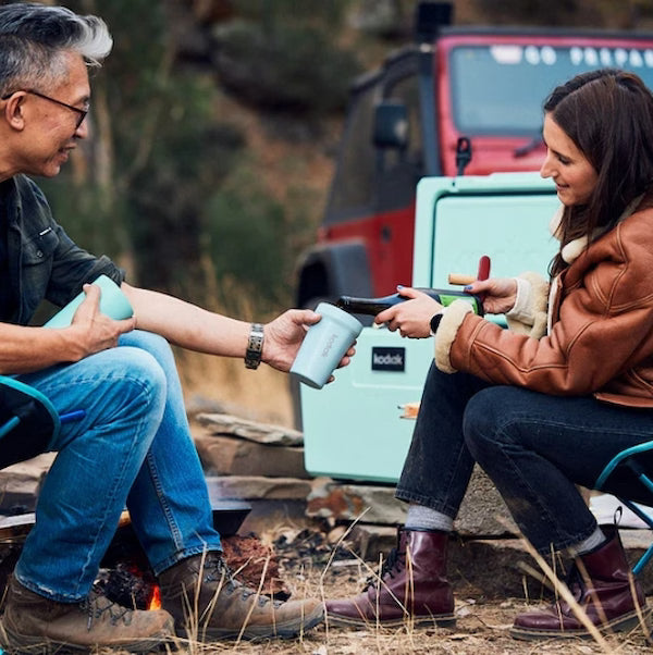 Two people sitting outdoors by a campfire, with a teal cooler and red vehicle in the background.