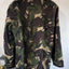 A military-style camouflage jacket with a high collar and shoulder epaulets, shown from the back against a plain off-white wall.