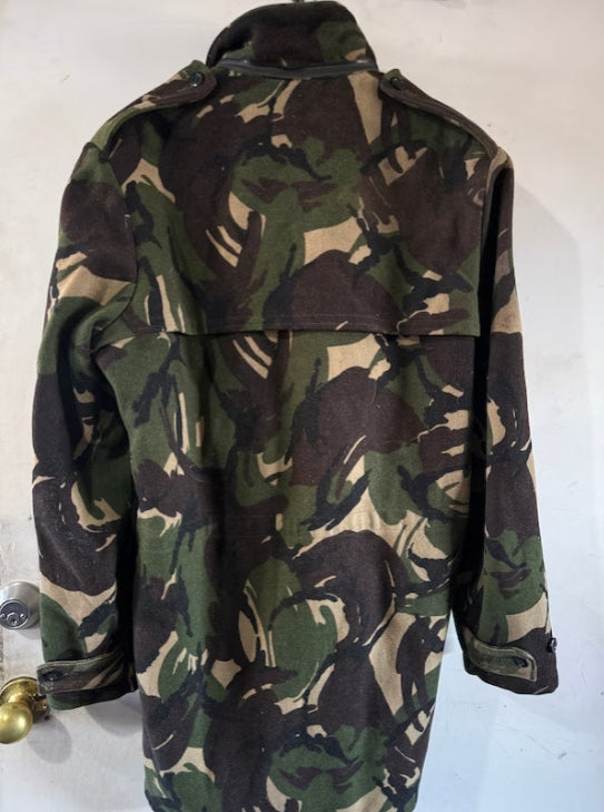 A military-style camouflage jacket with a high collar and shoulder epaulets, shown from the back against a plain off-white wall.