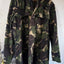 A woodland camouflage jacket hanging on a wooden hanger against a plain off-white wall.