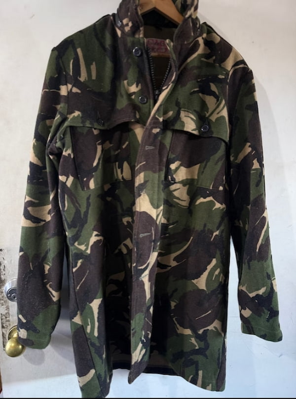 A woodland camouflage jacket hanging on a wooden hanger against a plain off-white wall.