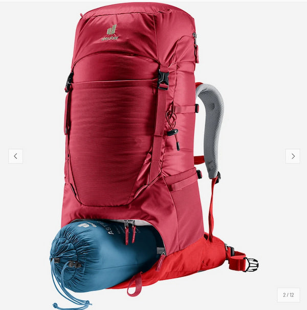 A red Deuter hiking backpack shown on a light gray background, with a blue sleeping bag tucked underneath. The backpack features multiple zippered compartments, side compression straps, and a gray padded shoulder strap.