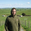 Man wearing a green hoodie standing on a dirt path with a scenic landscape of green fields and hills in the background.