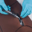 A close-up of a person wearing bright blue nitrile gloves applying Aquaseal waterproofing sealant to a seam on a brown waterproof suit or garment.