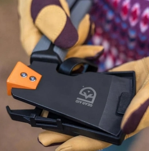 Black tool with orange clip held by a gloved hand, blurred colorful background
