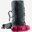 A dark gray Deuter hiking backpack with multiple compartments, shown with a pink trekking pole tucked into a side pocket and a water bottle in a side pouch, on a light gray background.