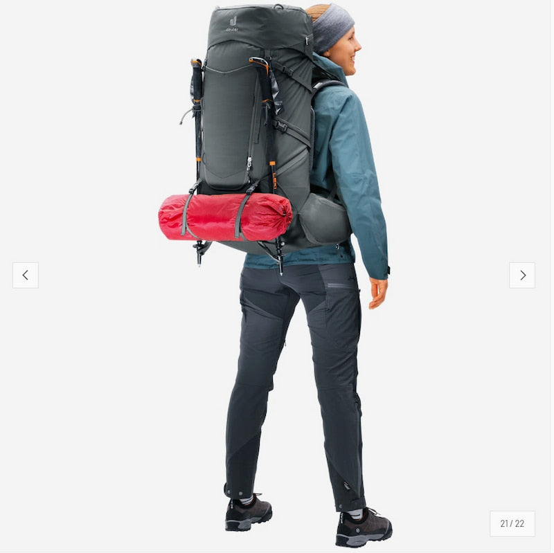 A person shown from the back wearing a large dark gray hiking backpack with a red sleeping bag or gear roll attached, a teal jacket, dark pants, and black hiking boots on a light gray background.