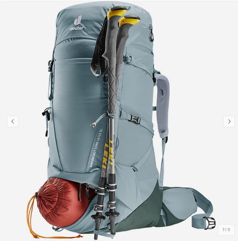 A Deuter hiking backpack in slate blue-gray, shown with two hiking poles and a red sleeping bag pouch against a light gray background.