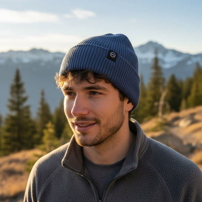 A man outdoors wearing a ribbed navy blue knit beanie with a small black circular logo patch and a gray fleece quarter-zip jacket, with mountains and trees in the background.