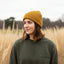 A woman wearing a mustard yellow ribbed knit beanie and an olive green crewneck sweatshirt standing in a field of dry golden grass.