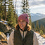 A woman outdoors in a snowy mountain landscape wearing a pink ribbed knit beanie with a small black logo patch and a dark olive green puffer vest over a cream fleece. Trees and rocks are visible in the background.