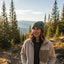 A woman wearing a beige sherpa fleece jacket and a green knit beanie stands outdoors in a mountain pine forest during golden hour.