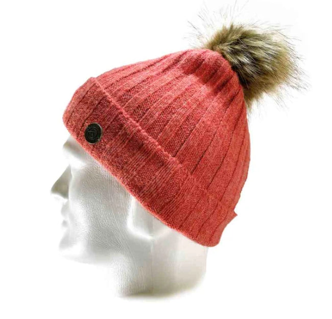 A ribbed knit beanie in a muted coral pink color, featuring a brown faux-fur pom-pom and a small black button on the side, displayed on a white mannequin head against a white background.