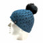A blue cable-knit beanie hat with a black faux-fur pom-pom and a small black logo patch on the side, displayed on a white mannequin head against a white background.