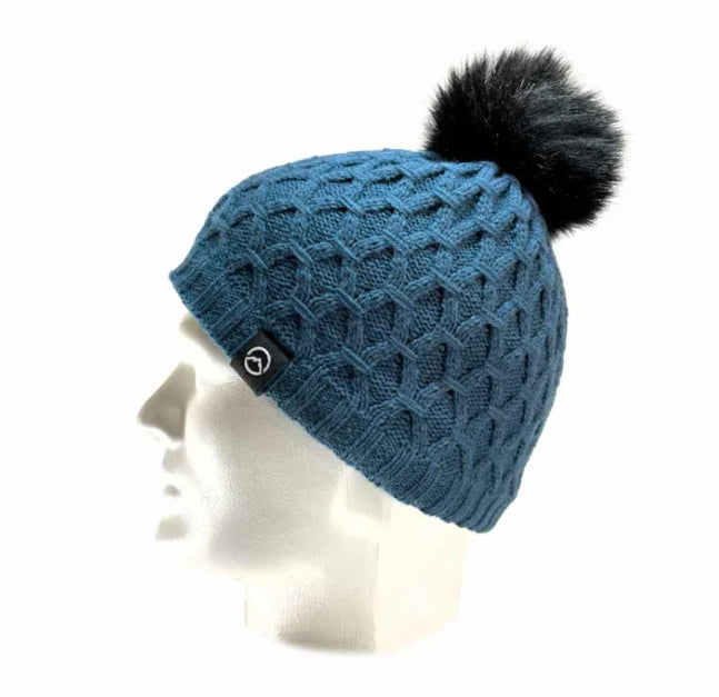 A blue cable-knit beanie hat with a black faux-fur pom-pom and a small black logo patch on the side, displayed on a white mannequin head against a white background.