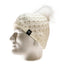 An off-white cable-knit beanie hat with a fluffy pom-pom on top, displayed on a white mannequin head. A small black square tag with a white circular logo is attached to the side of the hat.