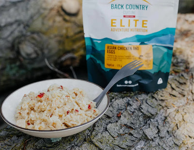 Bowl of food with a fork next to a Back Country Elite package on a rocky surface