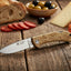 Knife with wooden handle on a wooden surface with bread, olives, and cheese in the background