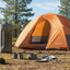 Orange dome tent with camping gear on a grassy field with trees in the background