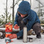 A person in a blue winter jacket crouches in the snow outdoors, assembling a red and gray camping stove next to a stainless steel thermos and a small cup.