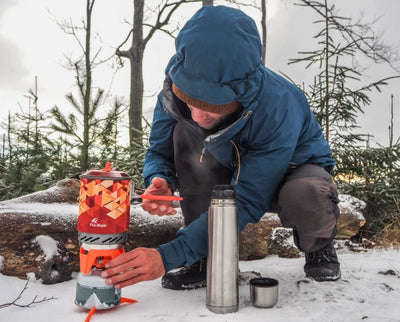 A person in a blue winter jacket crouches in the snow outdoors, assembling a red and gray camping stove next to a stainless steel thermos and a small cup.