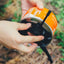 A close-up of a person's hands unscrewing a black plastic fuel cap from an orange and silver camping stove canister, set against a blurred outdoor background of green foliage and dry grass.