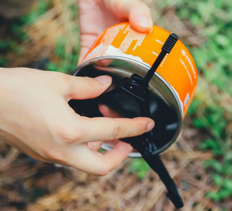 A close-up of a person's hands unscrewing a black plastic fuel cap from an orange and silver camping stove canister, set against a blurred outdoor background of green foliage and dry grass.