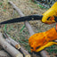 A black folding hand saw with the brand name 'FRENZALE' printed on the blade, held by a person wearing yellow leather work gloves while cutting a tree branch outdoors on dry grass and logs.