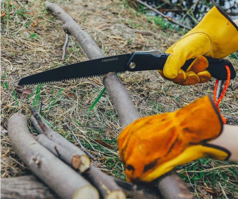 A black folding hand saw with the brand name 'FRENZALE' printed on the blade, held by a person wearing yellow leather work gloves while cutting a tree branch outdoors on dry grass and logs.