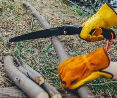 A black folding hand saw with the brand name 'FRENZALE' printed on the blade, held by a person wearing yellow leather work gloves while cutting a tree branch outdoors on dry grass and logs.