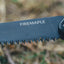 Close-up of a black Firemaple folding knife blade with serrated teeth, showing the brand name 'FIREMAPLE' in white text, set against a blurred outdoor background of dry grass and foliage.